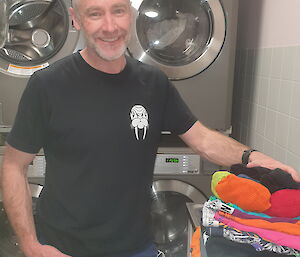A smiling man standing in front of four washing machines has just folded a pile of colourful clothes