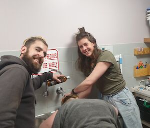 A smiling man and women wash a woman's hair as she leans over a laundry sink.