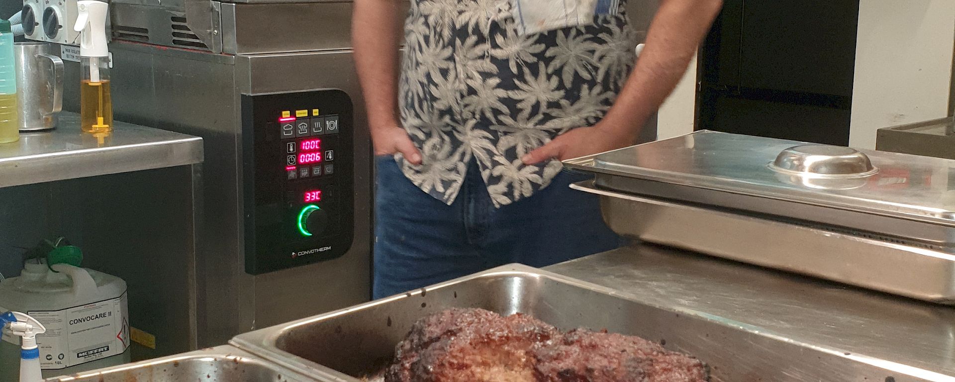 A smiling man wearing a fedora hat and a floral shirt with a tea-towel draped over his shoulder stands next to a kitchen bench on which sits two trays of cooked meat.