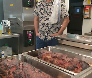 A smiling man wearing a fedora hat and a floral shirt with a tea-towel draped over his shoulder stands next to a kitchen bench on which sits two trays of cooked meat.