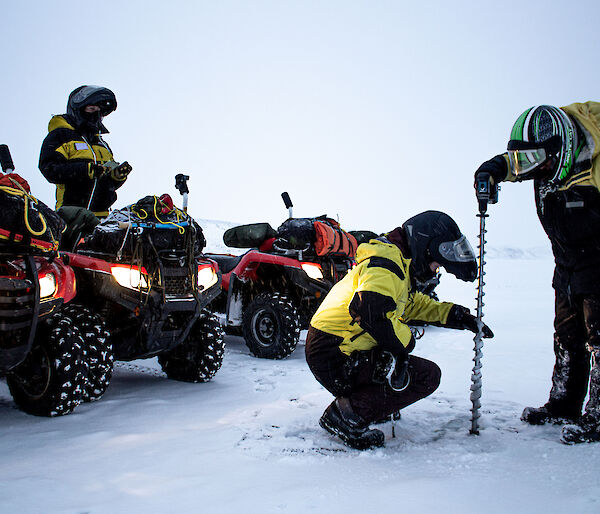 Two people wearing yellow snow gear using a very large drill to drill a hole into the sea-ice. There are three red quad bikes in the background and one person watching on.