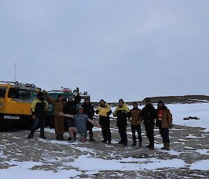 Ten people are in a line in front of three Hägglund vehicles on a rocky, snow and ice covered slope