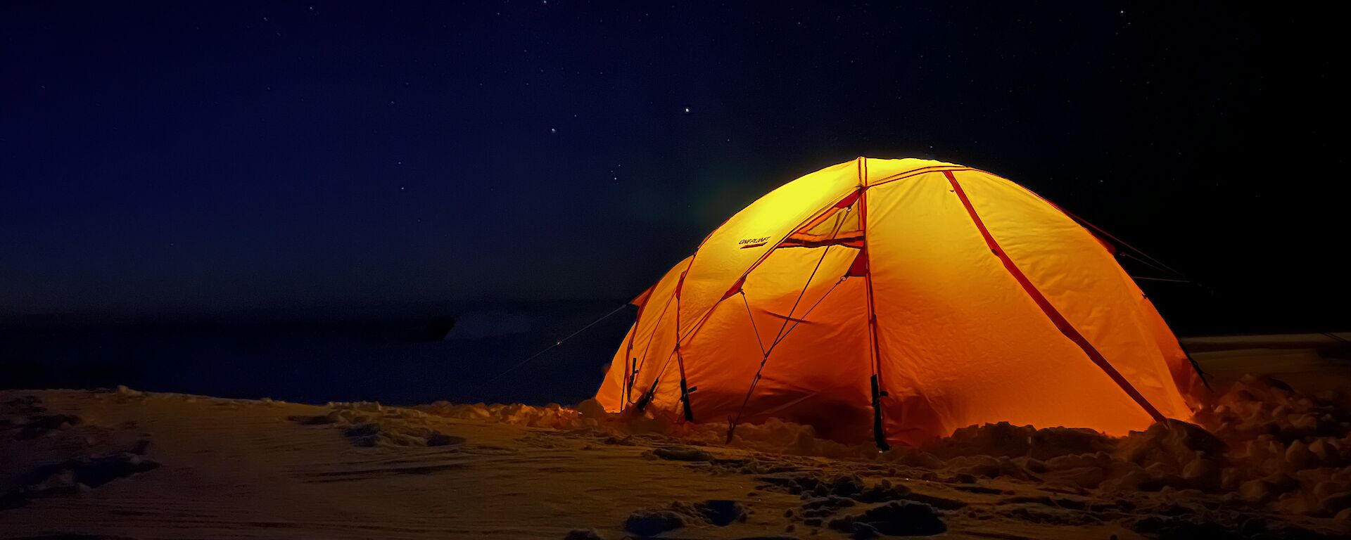 It is night time, and a yellow dome shaped tent sits on the snow covered ground. There is a light inside the tent, illuminating it. The sky is a very dark blue, almost black, and filled with stars.