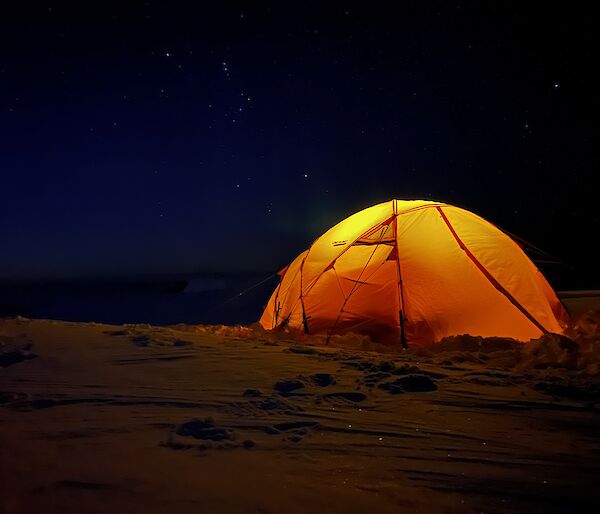 It is night time, and a yellow dome shaped tent sits on the snow covered ground. There is a light inside the tent, illuminating it. The sky is a very dark blue, almost black, and filled with stars.