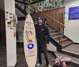A man in black jeans and top stands holding a wooden surfboard, and smiling. The surfboard has been painted with a picture of the man and a Happy Birthday message.