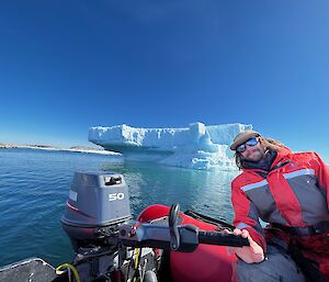 This photo shows the back section of a red inflatable boat, with a small outboard engine mounted to it. There is a man in view to the right of the frame who is wearing a red and grey survival suit, hat, and sunglasses. Behind him is a large white and blue iceberg, floating in the water. The sky is clear and blue.