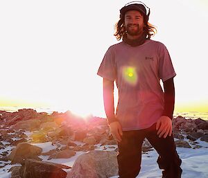 A man wearing a t-shirt and jeans stands facing the camera and smiling. The ground is snow covered, with exposed rock stretching into the background where the setting sun can be seen just above the horizon.