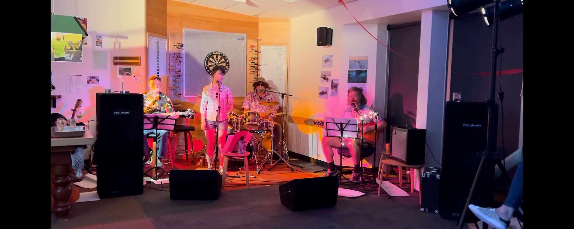 This photo is of the Casey band in action. From left to right there is a guitar player sitting, a lady standing at the microphone singing, the drummer in the background, and then another sitting guitar player. There is speaker equipment on the floor in front of the band.