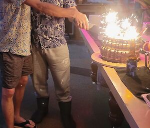 To the right of the picture is a large chocolate cake on a table, with several burning sparklers on it. Two men are to the left of the table and both holding the same knife, poised just above the cake and about to cut into it. Both men are smiling.