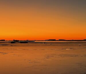 This photo is divided in two by the horizon. The top half is a range of orange shades, getting stronger towards the horizon. The foreground is a layer of sea ice, and large icebergs can be seen in the background. The sun has just set.