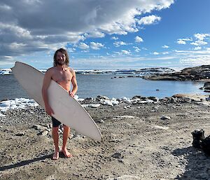 A man is standing holding a white surfboard under his arm and looking at the camera smiling. There is calm flat water in the background and small pieces of snow on the ground nearby. The sky is half filled with clouds.