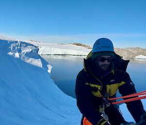 A man in a thick yellow and black jacket is being lowered down a snow covered hill, with calm blue water in the background. He has a bright orange rope attached to him, and he also has a blue helmet and sunglasses on.