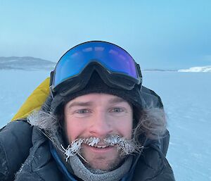 The frame is filled by a man looking at the camera with ice all through his moustache! He is dressed very warmly in a thick black jacket, beanie and large goggles. The background is a flat expanse of white sea ice, and a blurry low rocky hill.
