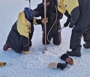 Three men leaning over and installing a cane into the sea-ice. A sensor string is attached to the cane.