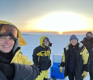 A team of four smiling men standing on the sea-ice with the sun sitting on the horizon in the background.