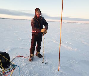 A smiling man holds a drill with a 1.5m drill bit while he drills the sea-ice.