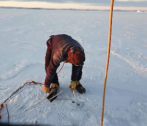 A man bending down and using a tape measure and string to measure how thick the sea-ice is using the hole he has just drilled.