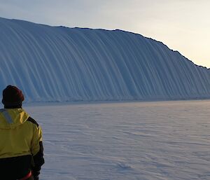 A man looks into the distance to a large ice-berg marked by spectacular vertical striations.