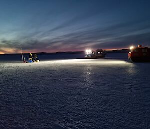 Two men, with their work area lit up by the headlights of vehicles, install scientific measuring equipment into the sea-ice.