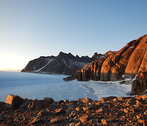 A landscape picture of rocky mountains rising from an ice plateau stretches into the distance with the sunset lighting the range