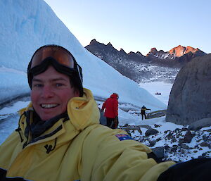A man is smiling into the camera with two people in the background and snow and ice rising to the side