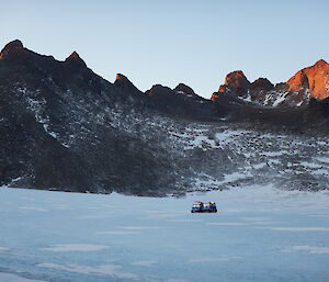 A blue vehicle is in the distance moving across the ice plateau with snow covered rocky peaks in the distance