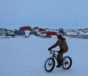 A man is riding a bicycle on the ice of a frozen harbour with station buildings in the background