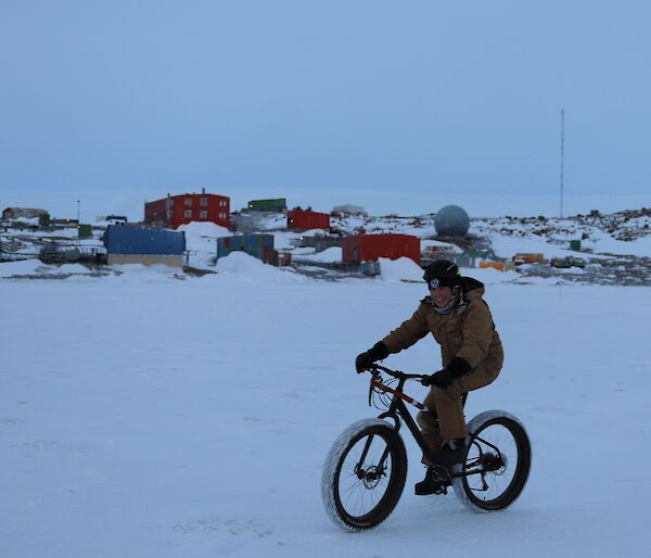 A man is riding a bicycle on the ice of a frozen harbour with station buildings in the background