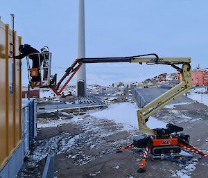 A man is on an elevated work platform repairing an antenna on the roof of a large yellow building