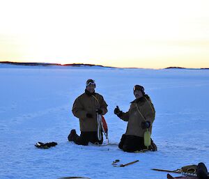 Two men are smiling at the camera while kneeling on an iced over harbour with the sun setting in the distance