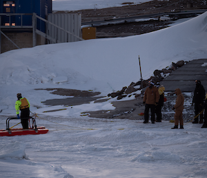 A man is standing on a rescue platform on top of an iced over harbour with a number of people watching from the shore