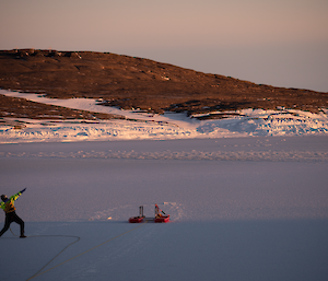 A man is standing on an iced over harbour pointing into the sky with a rocky hill rising in the background