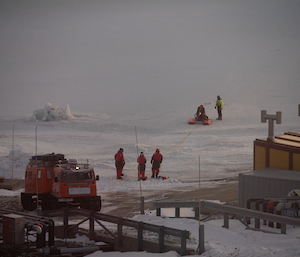 A team of people in immersion suits are conducted rescue training on an iced over harbour with a Hägglunds vehicle on the shore