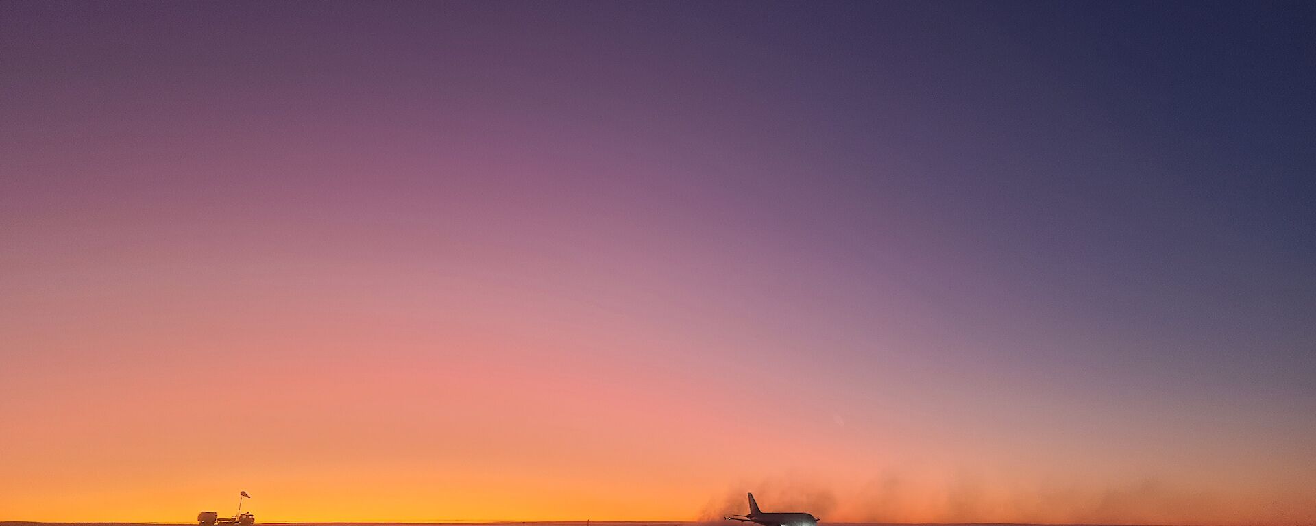 This photo is of the ice runway area at Wilkins Aerodrome at sunrise. There is a large white commercial aircraft in the lower middle of the picture, silhouetted with the orange and purple sky behind it. To the left of the photo is a small square vehicle.