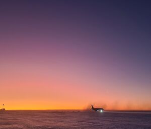 This photo is of the ice runway area at Wilkins Aerodrome at sunrise. There is a large white commercial aircraft in the lower middle of the picture, silhouetted with the orange and purple sky behind it. To the left of the photo is a small square vehicle.