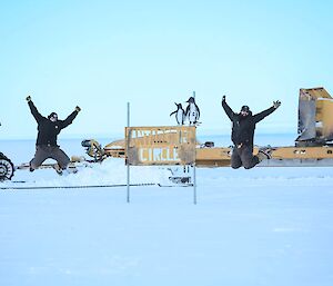 In the centre of this photo is a metal sign, with the words 'Antarctic Circle' cut out of it. Either side of this sign are two people caught in a mid air pose. The flat ground is covered in snow. To the right of the photo is the front blades of a yellow bulldozer, and to the left is the front of a red tractor.