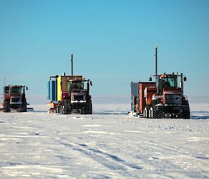 The photo is divided in two horizontally, with snow in the lower half and clear blue sky in the upper half. There are three large tractors heading in line towards the camera, all towing big rectangular containers.