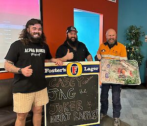 Three men stand behind a chalkboard with 'Jag the Joker + Meat Raffle' written on it. The man on the right is holding a large tray of various meats covered in plastic wrap. All three men are smiling with their thumbs up.
