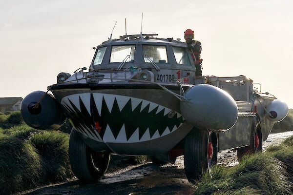An amphibious vehicle travels down a wet road surrounded by grassy tussocks