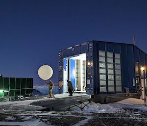 Two men in protective suits are exiting a blue building through a large door. The man in the lead is holding a large helium-filled weather balloon ready for release