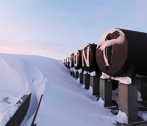 A large drift of snow is to the left of a number of fuel tanks which are raised above the ground on concrete stands