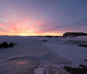 An aircraft hangar is to the right of frame with the sun setting in the distance over an ice covered harbour