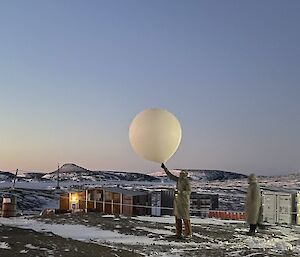 A man in a protective suit is releasing a large helium filled weather balloon, watched on by another man in a protective suit