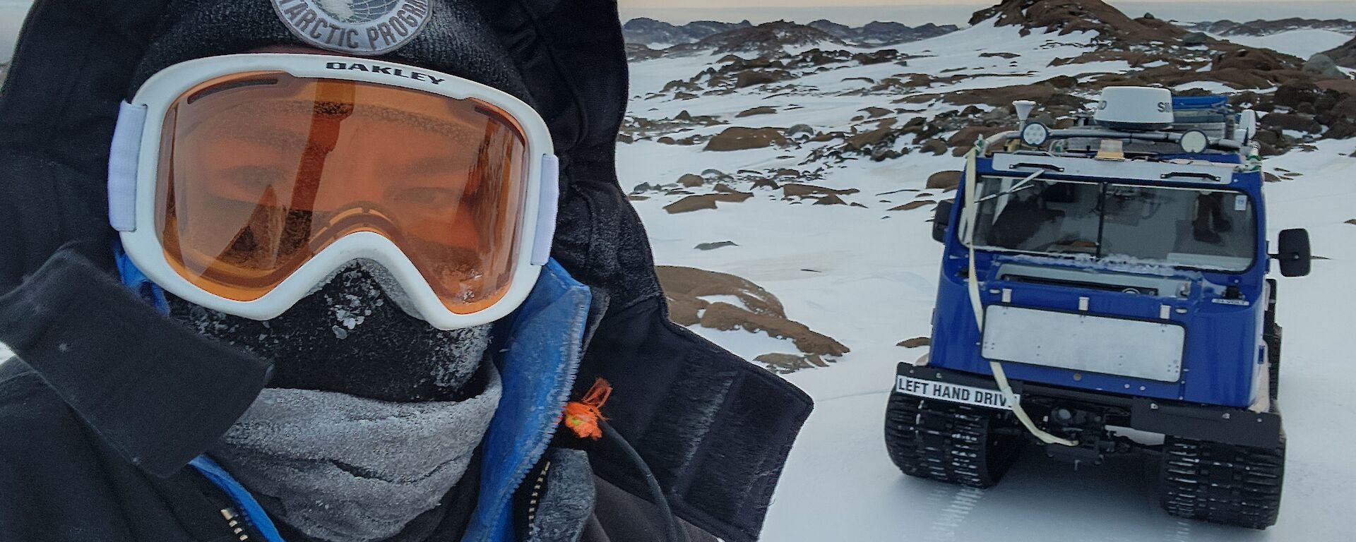 A man stands to the left of the photo, close to the camera and filling half of the shot. He is wearing a big black jacket with a hood, with goggles and a full face covering. In the background on the right is a snow covered hill with a square blue tracked vehicle parked with it's front facing the camera.