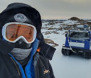 A man stands to the left of the photo, close to the camera and filling half of the shot. He is wearing a big black jacket with a hood, with goggles and a full face covering. In the background on the right is a snow covered hill with a square blue tracked vehicle parked with it's front facing the camera.