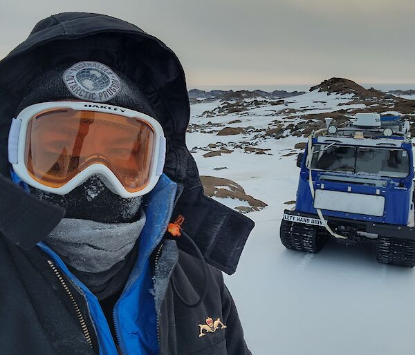 A man stands to the left of the photo, close to the camera and filling half of the shot. He is wearing a big black jacket with a hood, with goggles and a full face covering. In the background on the right is a snow covered hill with a square blue tracked vehicle parked with it's front facing the camera.