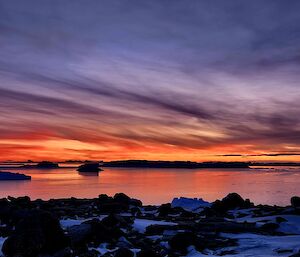 The top half of the photo is a sky filled with streaks of purple and orange cloud. Below this, the colour is reflected in flat sea ice that leads down to a snow and rock covered shoreline.