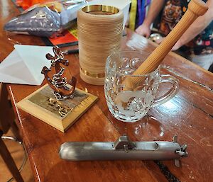 Sitting on a wooden bar top are some birthday gifts. A stubby cooler turned up from plywood used in a C-17 airdrop, hand carved wooden anchor, a steel submarine shaped bottle opener, and an etched glass with a handle. Inside the glass sits a miniature wooden baseball bat.