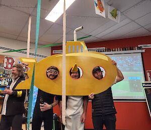 Three men hold up a yellow submarine cut out of plywood. The submarine has three holes cut in it, that frame the men's faces. The man in the middle is wearing a penguin costume! In the background is a large projector screen displaying a baseball game. The photo is taken in the bar and there are decorations like streamers hanging from the ceiling.