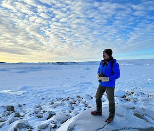 A man in a blue jacket, gloves and beanie, is standing to the right of frame atop a snow covered rock. He is staring off to the left of the photo and holding a camera with both hands in front of him. The snow covered sea ice extends into the background, and light puffs of cloud cover the sky in a blanket.